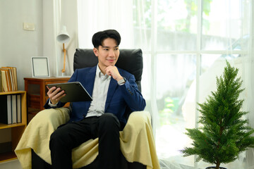 Confident businessman sitting in an armchair holding digital tablet and looking thoughtful