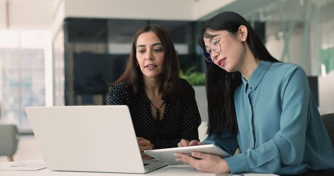Two young multiethnic product managers using gadgets at workplace, comparing marketing statistic data on tablet and laptop, analyzing online content, report, discussing Internet sales