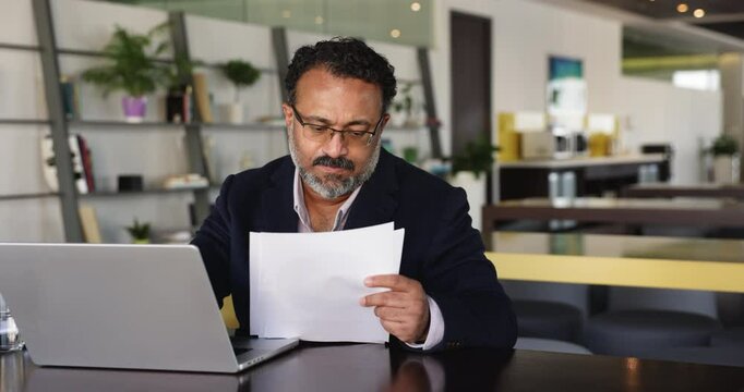 Serious senior Indian business owner man analyzing paper financial report, using online bank app, service, for paying bills, taxes on laptop, doing accounting work, paperwork, reading legal document
