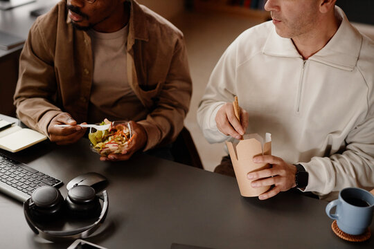 Cropped shot of man enjoying wok noodles having lunch together with colleague while watching at laptop screen in office