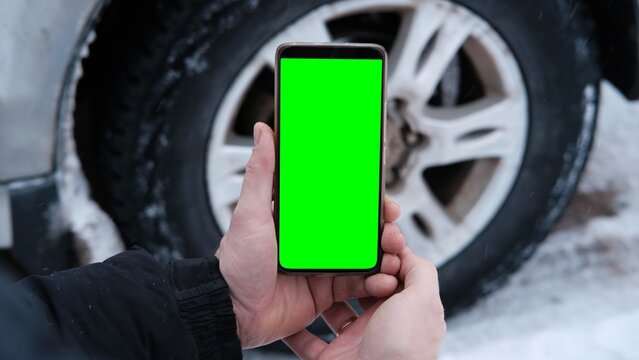 Male hands holding a smartphone with a bright green screen against the background of a car wheel. Automotive technology. Mobile applications for drivers. Road safety. Using gadgets. Chroma key - Powered by Adobe