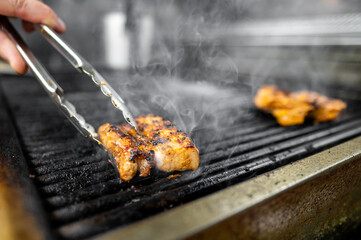 A hand using tongs to flip juicy, grilled meat on a barbecue grill, with steam rising and a smoky atmosphere, showcasing delicious cooking and outdoor dining.