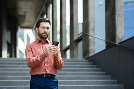 Serious thinking businessman with phone in hands reads online messages and browses internet pages. Man uses smartphone outside office building, walking through city.