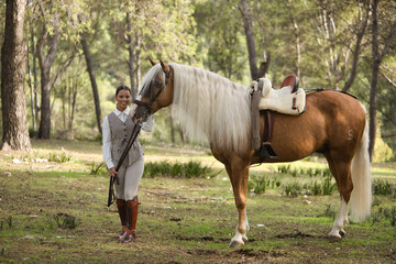 Young woman dressed as a horsewoman, smiling and happy looking at camera next to her beautiful brown horse with white mane, in the middle of a forest. Concept animals, horse riding, equestrian, horse,