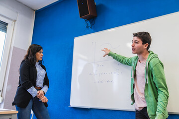 High school student explaining math formula at whiteboard to teacher