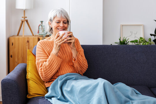 Portrait of mature adult woman smelling aroma coffee wrapped in a warm blanket in the sofa. Warm winter situations at home