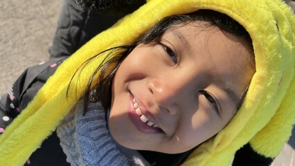 Immersive POV of close up funny smiling Six years old Indonesian girl standing and looking at camera using yellow beanie