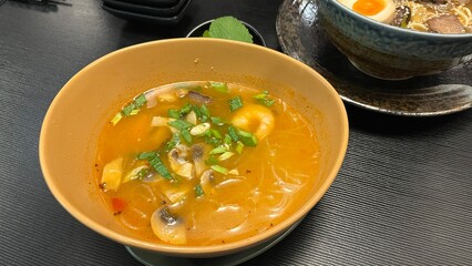 A bowl of Tom Yum soup, a traditional Thai hot and sour soup. Served in a rustic orange bowl on a dark textured table. Authentic Thai cuisine, healthy eating, and flavorful dining concept