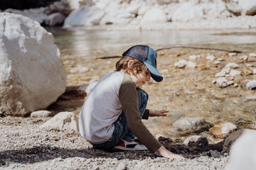 Caucasian school boy sits on a riverside rock in the canyon with mountain cliffs in the background. Kid child taking a rest on a boulder near mountain river bank.