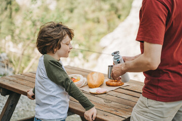 Father dad pours hot coffee tea from thermos into the mug on a family picnic in the mountains. Child school boy kid is watching his dad filling the cup with hot beverage.