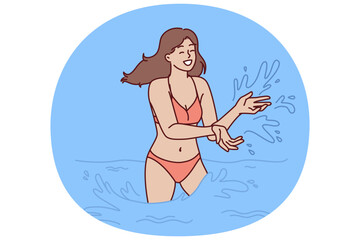 Woman in bikini swims in sea and splashes with water enjoying summer travel or trip to aquapark. Happy girl in swimsuit stands knee-deep in sea during vacation on tropical island with hot climate