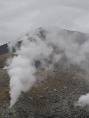a volcanic landscape with steaming fumaroles or vents emitting gases.  The terrain is rocky and barren, with a mountain slope rising in the background under a cloudy sky.
