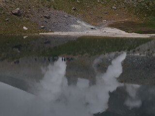 a volcanic landscape with steaming fumaroles or vents emitting gases.  The terrain is rocky and barren, with a mountain slope rising in the background under a cloudy sky.