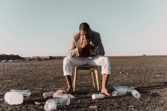 Young man sitting on chair surrounded by plastic bottles in barren land
