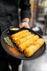 A chef holds a black plate with crispy golden fried sticks, garnished with thyme and a sprinkle of spice, served with a small bowl of dipping sauce.
