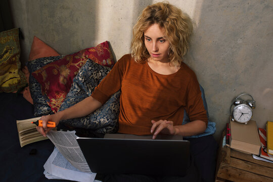 Portrait of blond student sitting on bed working on laptop