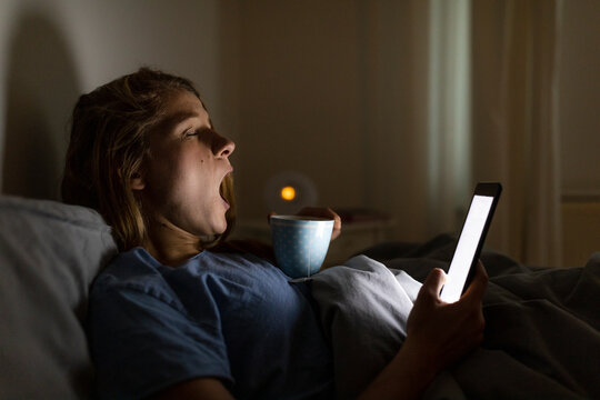 Young woman lying in bed at home with cup of tea at night using tablet