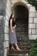 A woman stands on stone steps in an arch. She has long dark hair and is wearing a black top and...