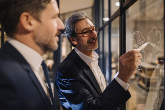 Two businessmen working on drawing on glass pane in office