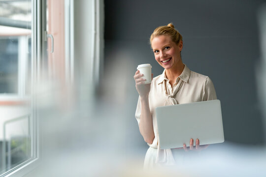 Smiling young businesswoman with laptop and takeaway coffee - Powered by Adobe