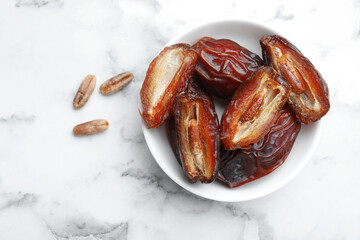 Many tasty dried dates with seeds in bowl on white marble table, top view