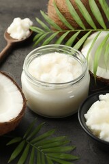 Organic coconut cooking oil in jar, spoon, fresh fruits and green branches on grey table, closeup
