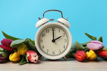 Spring time. Alarm clock and beautiful tulips on wooden table against light blue background