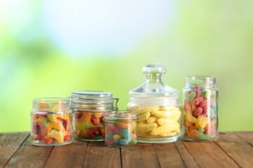 Tasty gummy candies in jars on wooden table against blurred green background