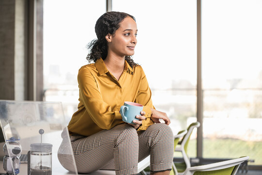 Young businesswoman sitting on desk in office having a coffee break - Powered by Adobe