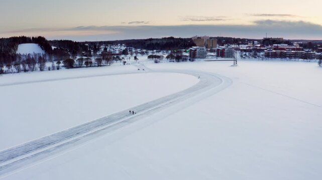 Drone Footage of Frozen Lake with Winding Ice Track Leads Toward a Snow-Covered Cityscape at Sunset, People Enjoying Winter Activities in Cold Winter Landscape in Pite&aring;, northern Sweden