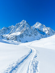Serene winter landscape: A snow-covered path winds through pristine snow towards majestic snow-capped mountains under a vibrant blue sky.