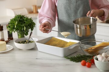 Woman spreading bechamel sauce onto spinach lasagna at marble table indoors, closeup
