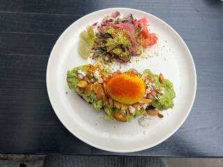 Avocado toast with a turmeric-poached egg, nuts, and a colorful side salad on a minimalist white plate. The vibrant greens and reds pop against the dark table.