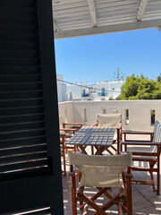 A shaded view through blue shutters onto a sunlit terrace with wooden chairs. The contrast of light and shadow captures a peaceful, breezy atmosphere.