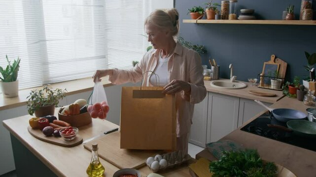 Medium shot of adult Caucasian woman unpacking fresh produce, including zucchini and tomatoes, from brown paper bag in sunlit kitchen with plants, wooden counters, and well arranged cooking essentials