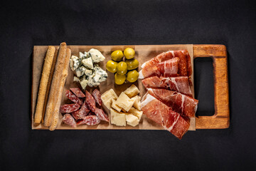 Gourmet charcuterie board with cheese, olives, cured meats, and breadsticks on a wooden tray against a black chalkboard background