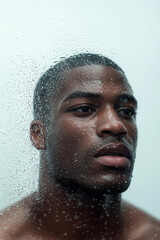 Close-up of a Black man looking through a wet glass window, deep in thought
