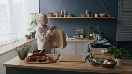 Medium shot of senior Caucasian woman organizing items from brown paper bag in modern kitchen filled with natural light, plants, and fresh produce, creating warm and homely atmosphere - Powered by Adobe
