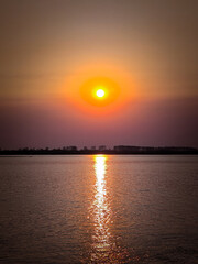 Naklejka premium The sun sets on the horizon, casting a golden and orange glow over a calm river. silhouetted trees line the riverbank, enhancing the tranquil and serene atmosphere. the reflection of the sun creates a