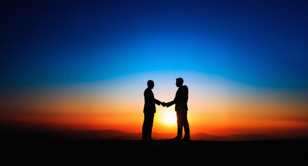 Two silhouetted figures shake hands against a striking sunset backdrop. The sky blends from deep blue to vibrant orange and red as the sun dips below the horizon, creating a symbolic scene of agreemen