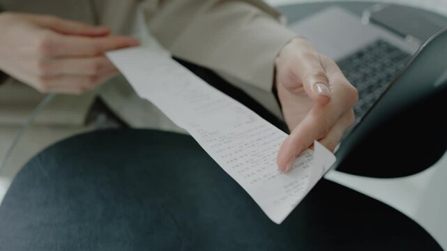 A close-up image showing a hand holding a receipt near a laptop in an office setting, symbolizing finance, accounting, and business transactions. Business Woman Checking Financial Receipt