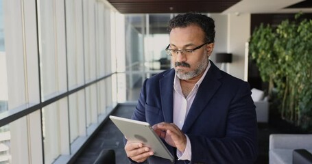 Serious senior Indian company CEO man using financial application on tablet, analyzing reports, stock market trading data, investment income, turning look away, touching chin, thinking - Powered by Adobe