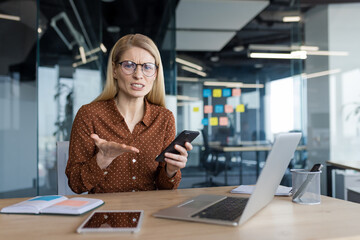Nervous upset woman at workplace in office looking at camera with smartphone in hands. Businesswoman received bad news online, and app crash freezes on phone.