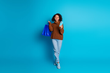 Confident young woman holding shopping bags and phone on vibrant blue background