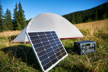 Tourist tent with solar panel and portable power station nearby in grassy field. Scene surrounded...