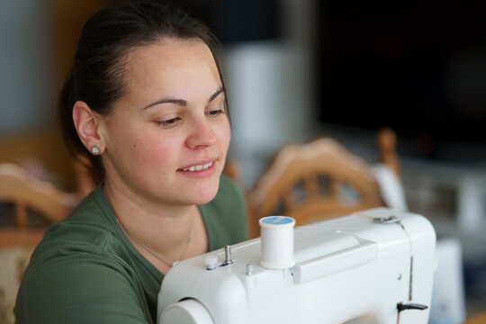 Cute, focused girl in a T-shirt sewing on a machine in a cozy room with a blurred background. Concept of home creativity, learning by doing, and childhood skills development.