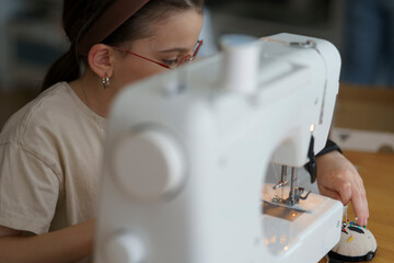 A little cute girl wearing glasses sitting at a sewing machine at a wooden table in a room. Educational and creative children's concept.