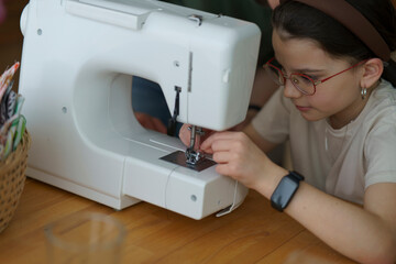 A little cute girl wearing glasses sitting at a sewing machine at a wooden table in a room. Educational and creative children's concept.