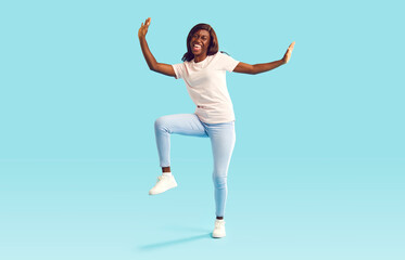Portrait of a cool funny young african american girl with joyful face expression dancing wearing casual clothes isolated on blue turquoise background. Happy excited woman having fun in studio.