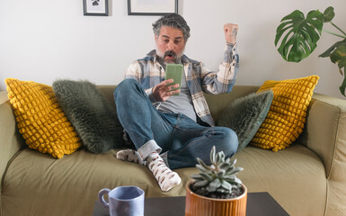 Happy man raising fist while looking at smartphone and celebrating success on sofa at home